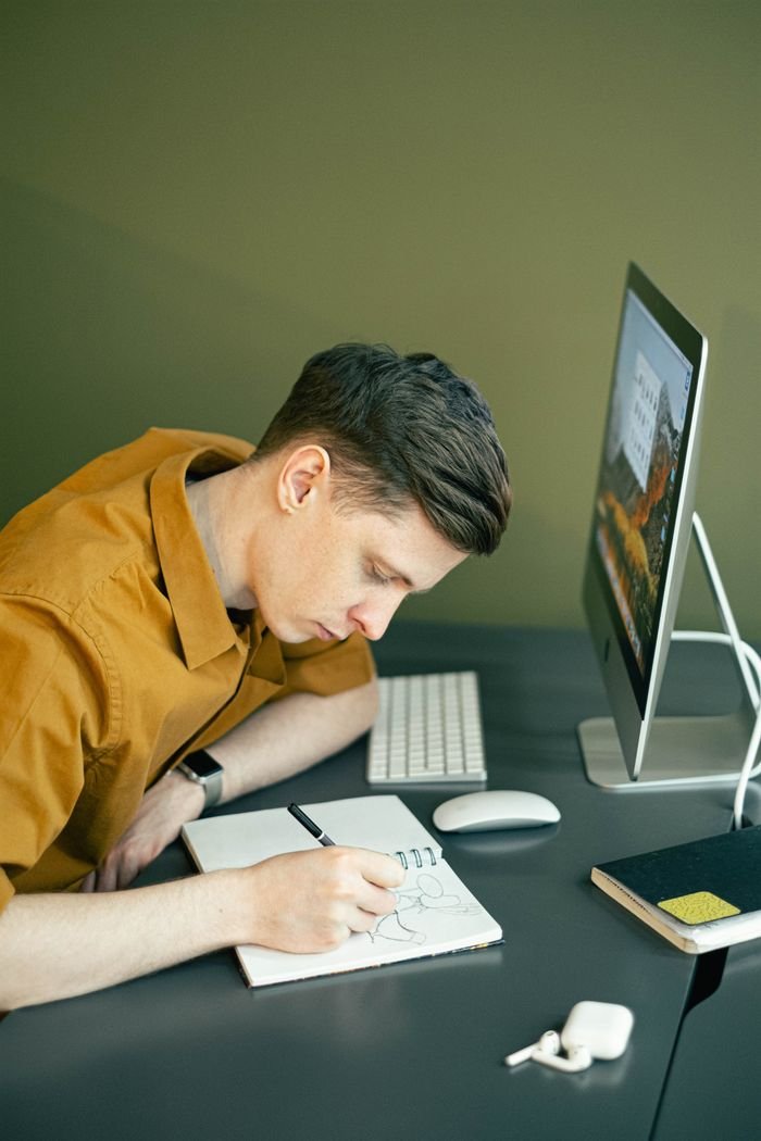 services-02 Young man sketching on paper at a desk with a computer, illustrating creative concentration.